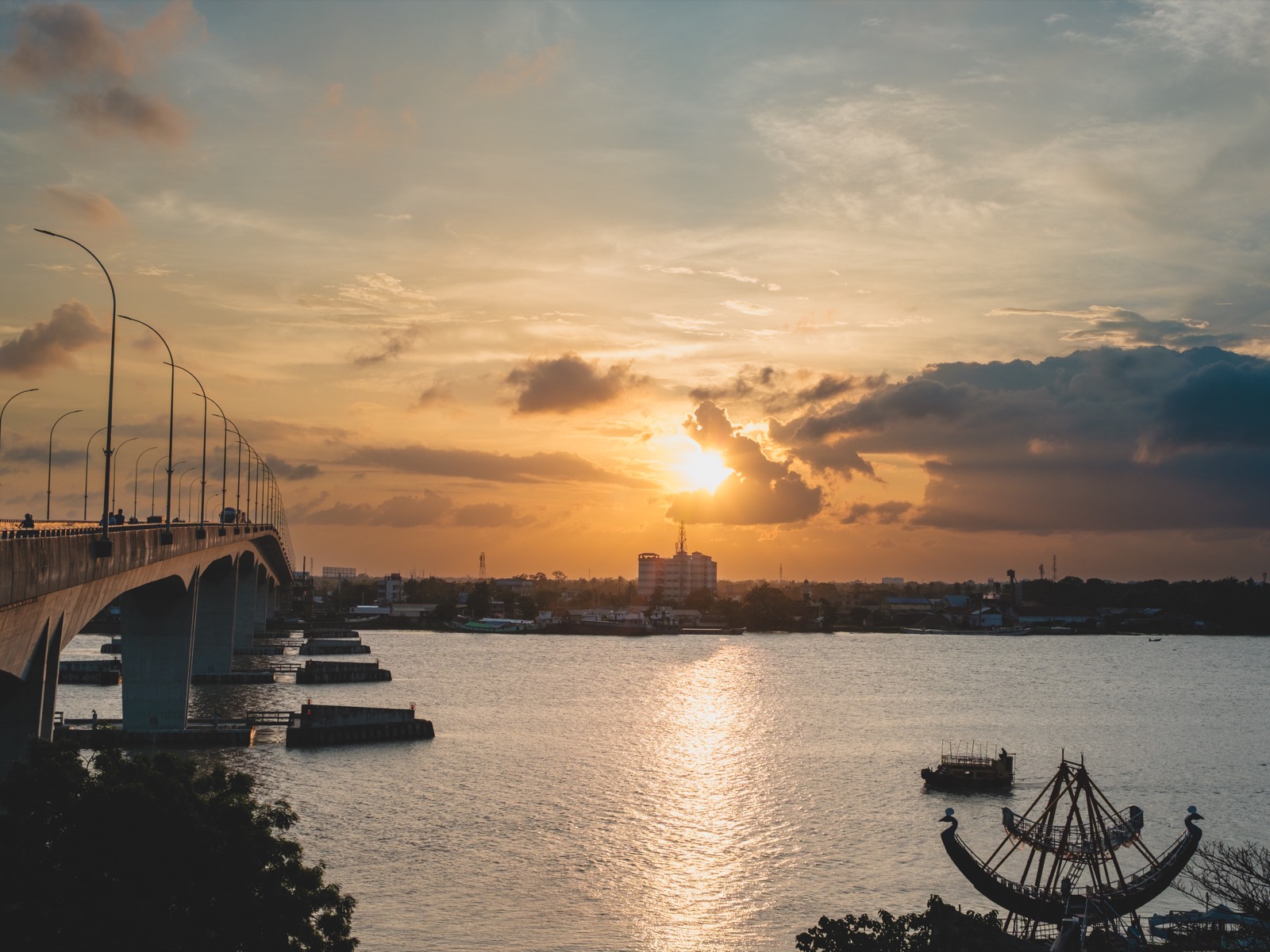A concrete bridge over a wide river at sunset with the sun low in the frame behind cloud, warm orange light reflecting off the water.