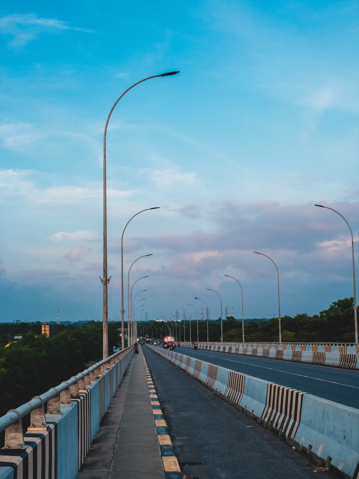 A long bridge walkway running to the horizon under a blue sky, lined with tall curved street lamps, shot at f/8 on the Voigtländer Ultron 27mm.