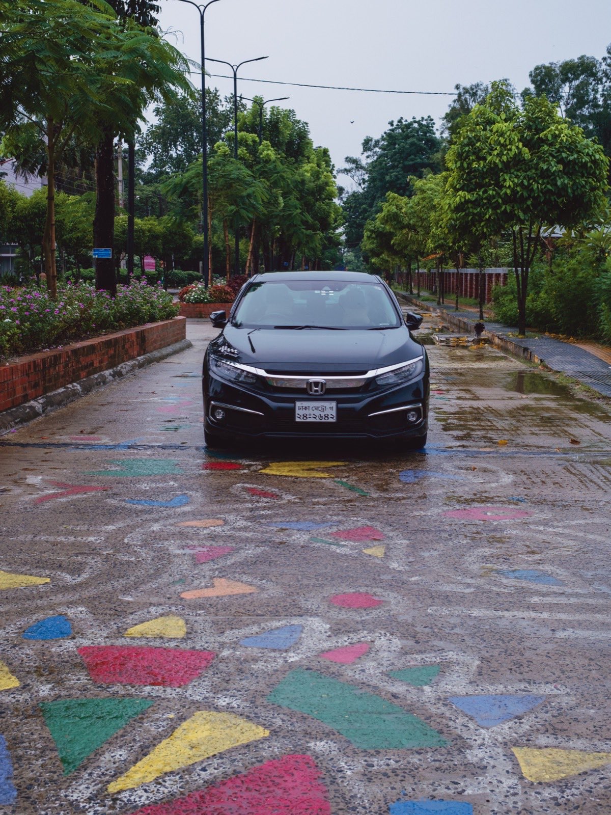 A black Honda Civic parked on a wet road painted with coloured geometric shapes, trees and flowers lining both sides, shot on the Voigtländer Ultron 27mm.