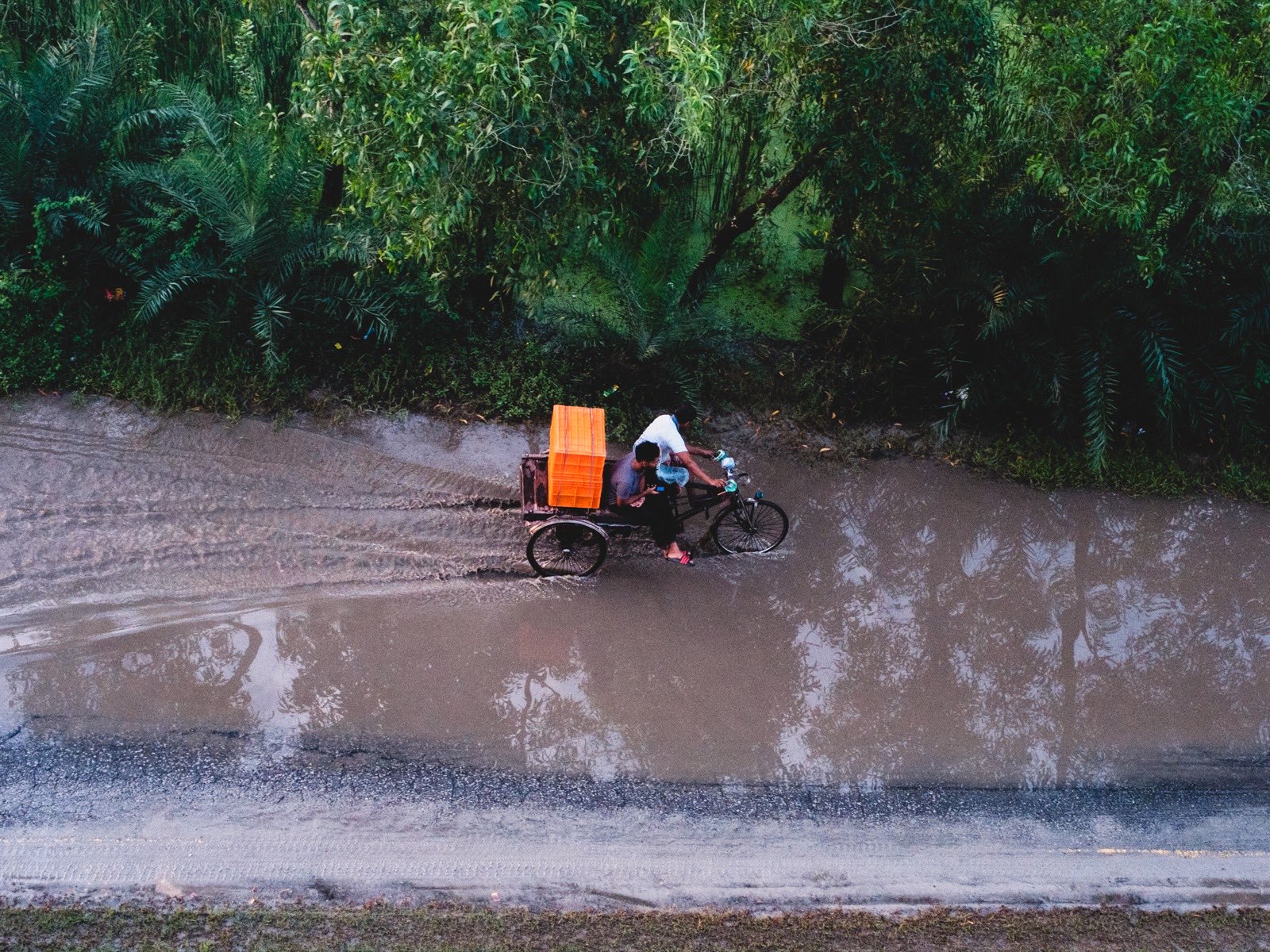 Overhead view of two men on a cycle rickshaw van loaded with orange crates, pushing through a flooded muddy road beside dense green trees.