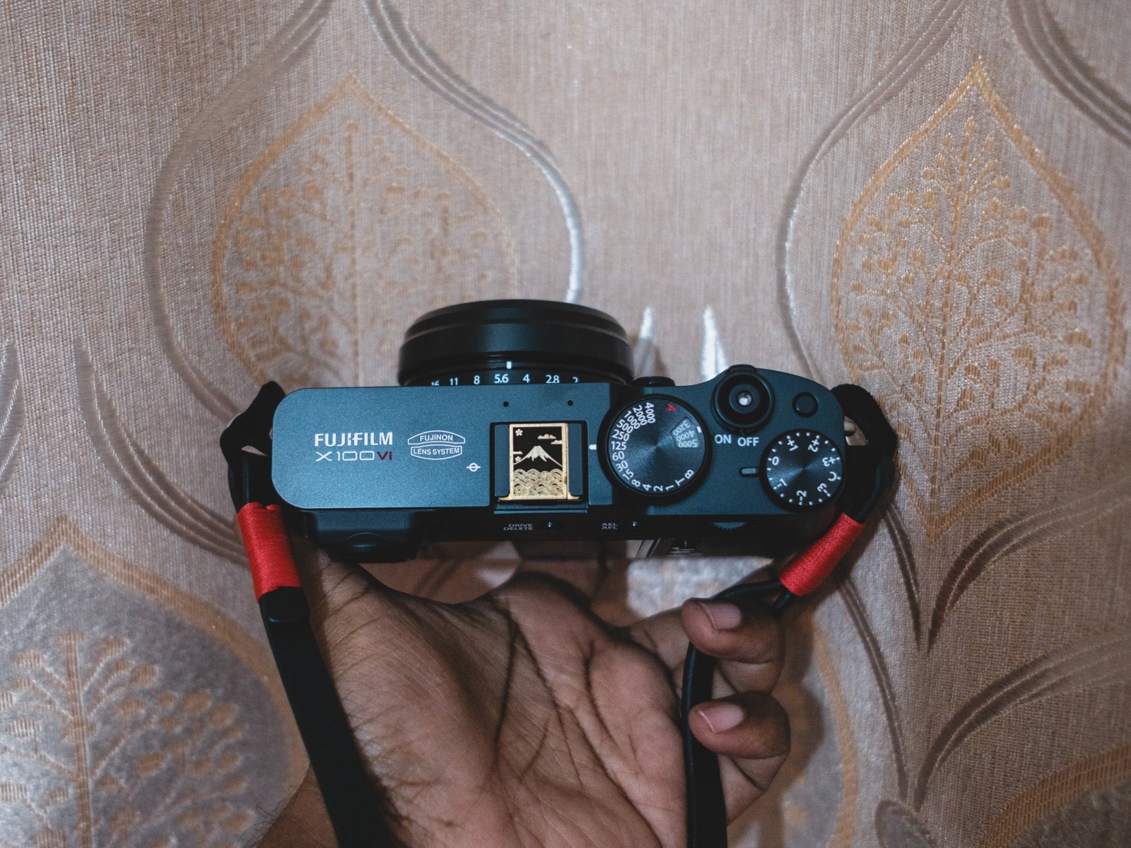 The top plate of a black Fujifilm X100VI held in one hand, showing the shutter speed dial, the exposure compensation dial and a Mount Fuji sticker over the hot shoe.